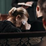 A grieving woman wipes tears during a heartfelt funeral ceremony in a somber indoor setting.